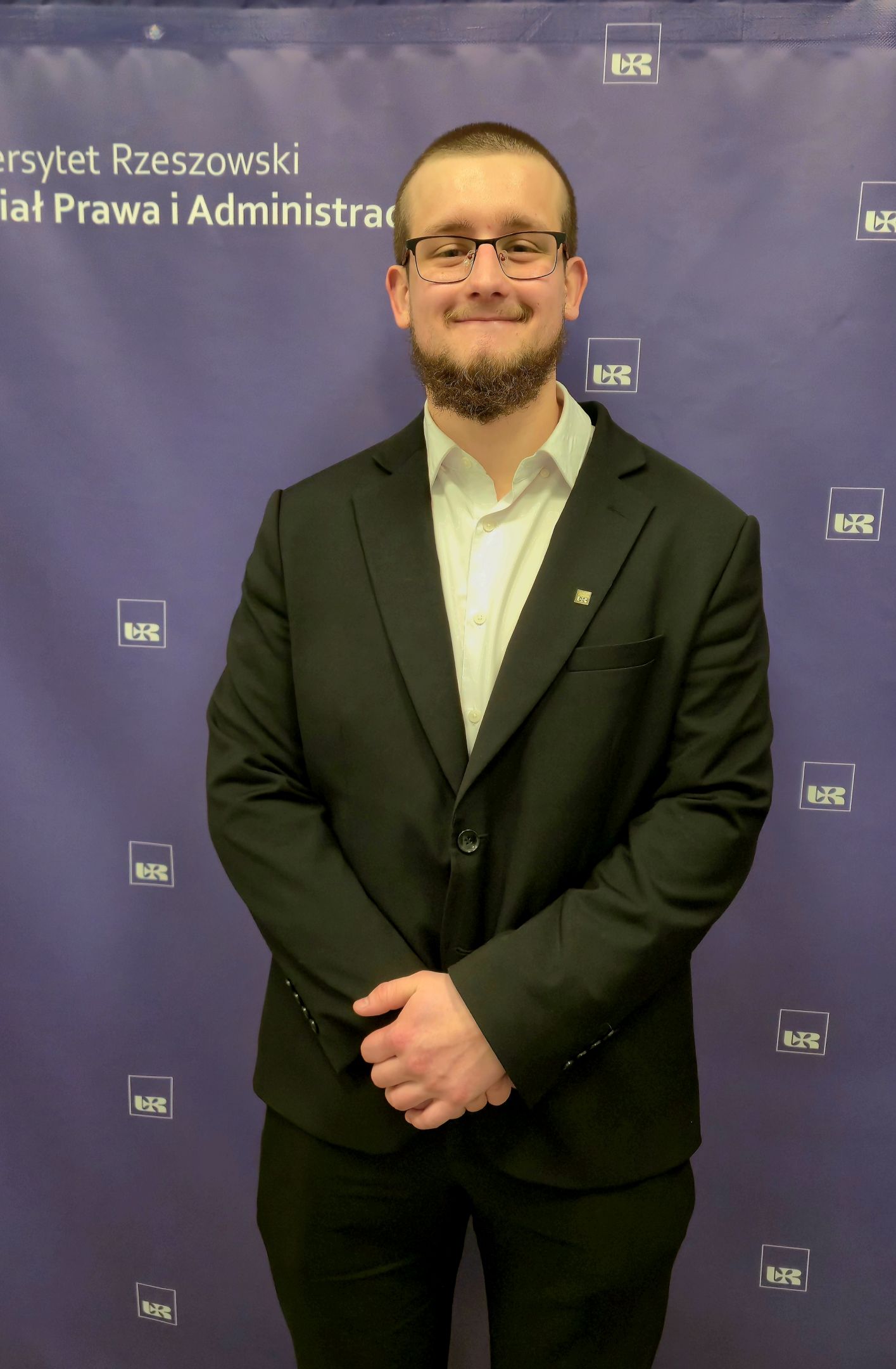 Mężczyzna ubrany w garnitur i koszulę, na tle z logo Uniwersytetu Rzeszowskiego i Wydziału Prawa i Administracji