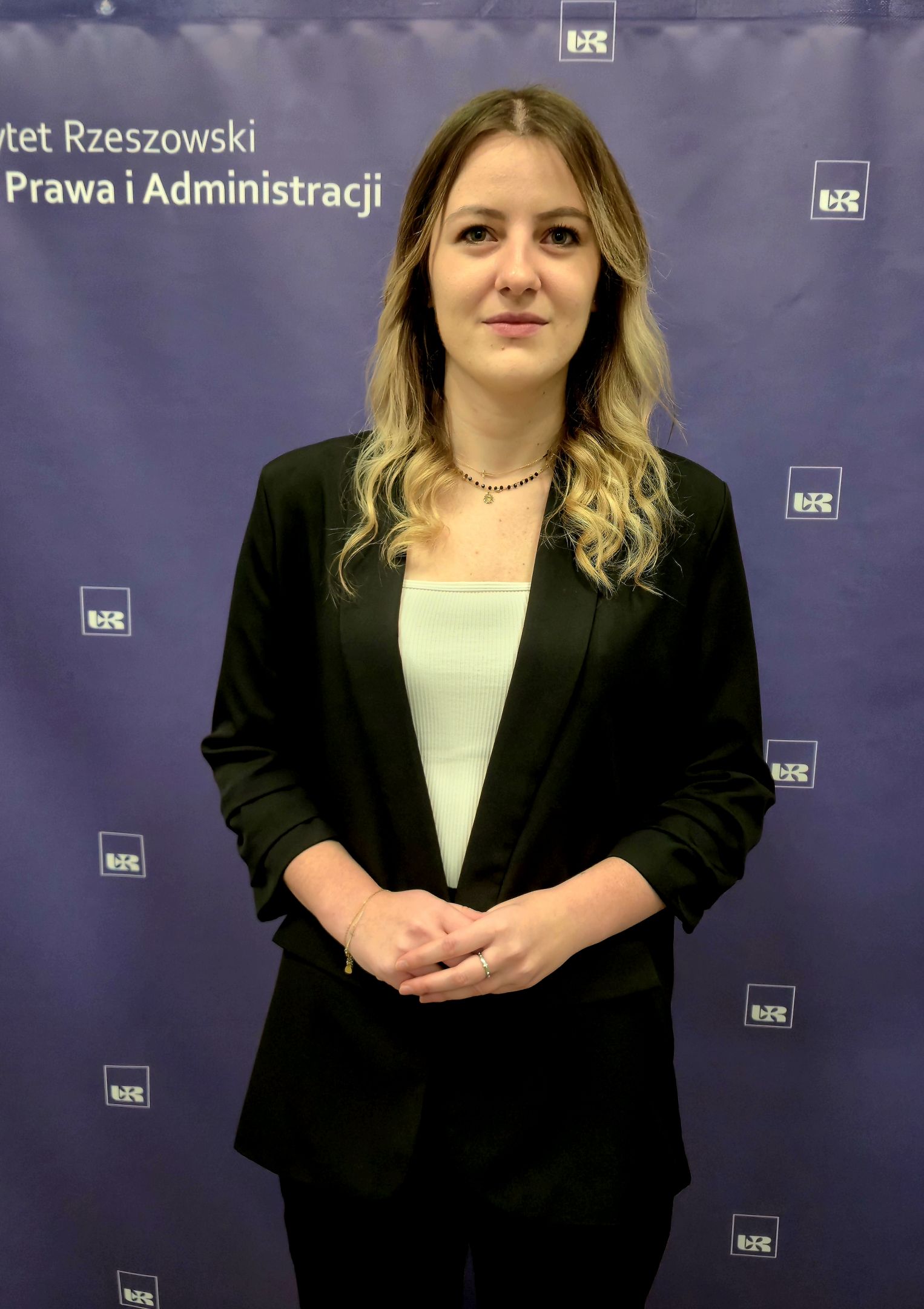 Kobieta ubrana w marynarkę i koszulę, na tle z logo Uniwersytetu Rzeszowskiego i Wydziału Prawa i Administracji