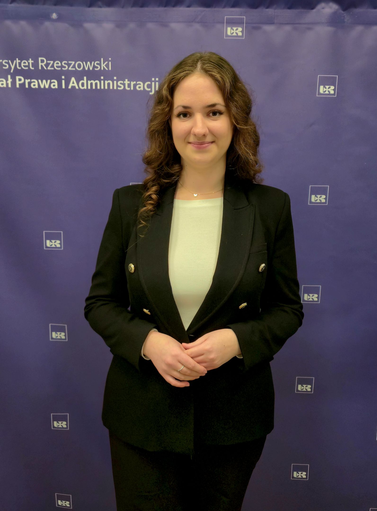Kobieta o ciemnych kręconych włosach ubrana w marynarkę i koszulę, na tle z logo Uniwersytetu Rzeszowskiego i Wydziału Prawa i Administracji