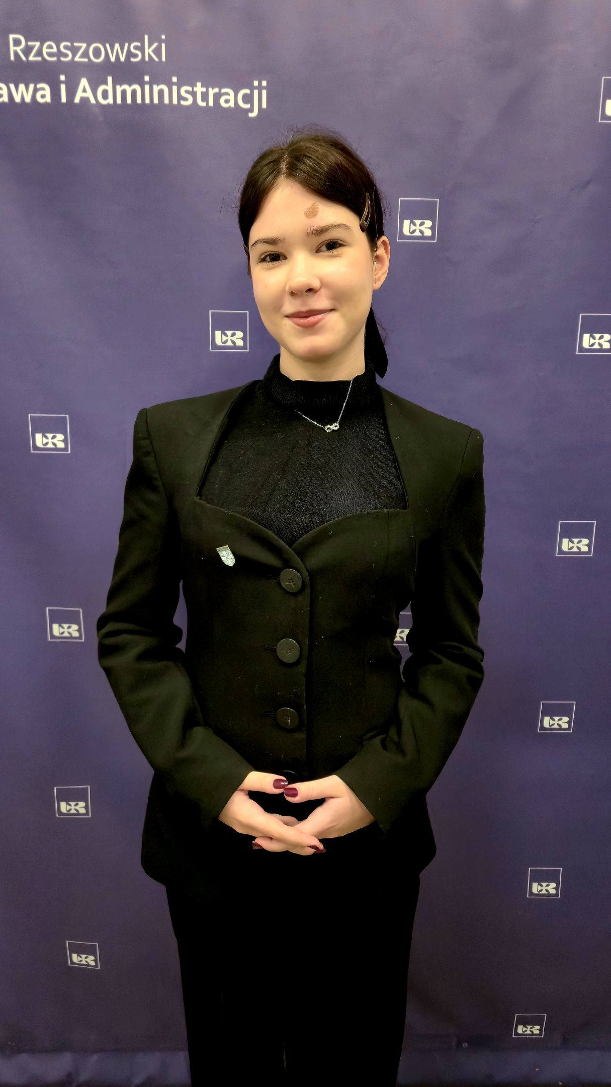 Kobieta o ciemnych upiętych włosach ubrana w marynarkę i koszulę, na tle z logo Uniwersytetu Rzeszowskiego i Wydziału Prawa i Administracji