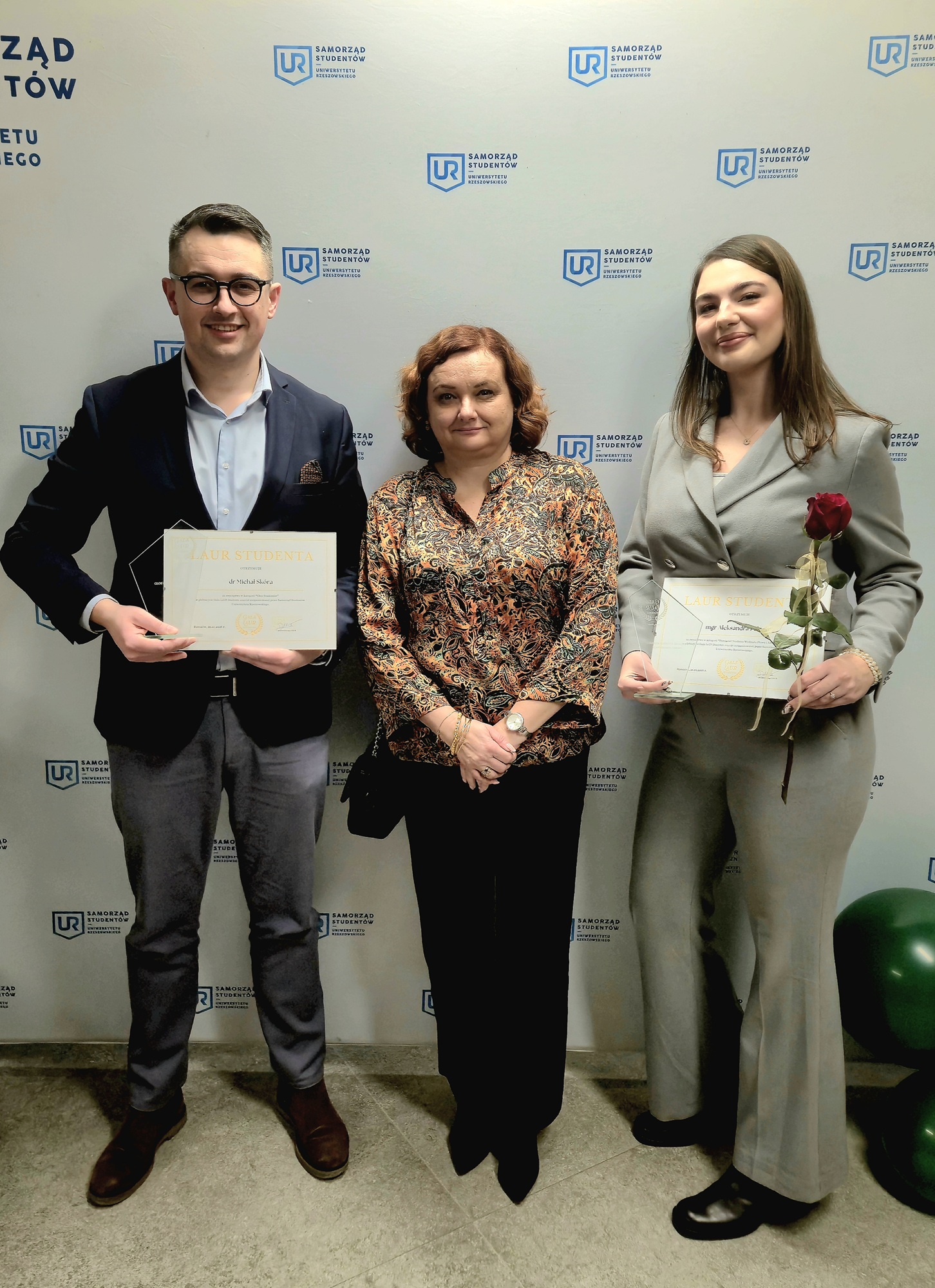 Trzy osoby (mężczyzna i dwie kobiety) stojące obok siebie, ubrane w stroje p&oacute;łformalne, trzymające w rękach dyplomy