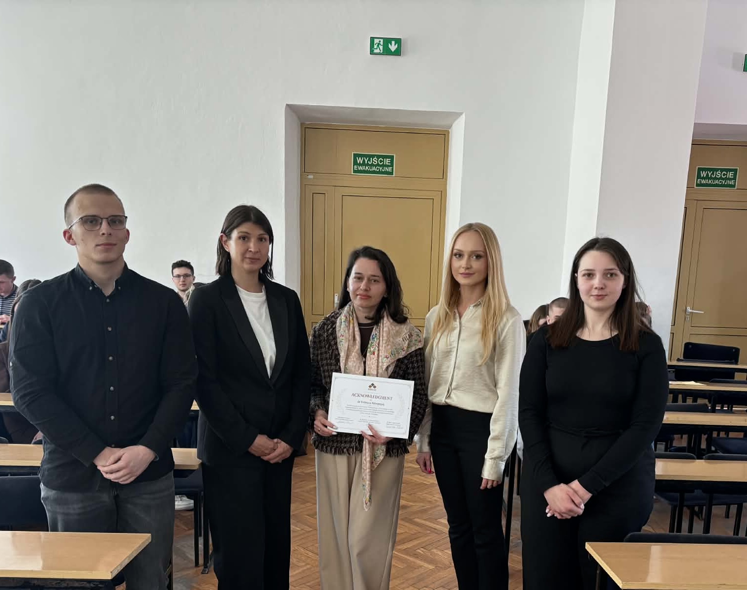 Grupa pięciu os&oacute;b (trzech kobiet i dw&oacute;ch mężczyzn) stojących w rzędzie w sali wykładowej